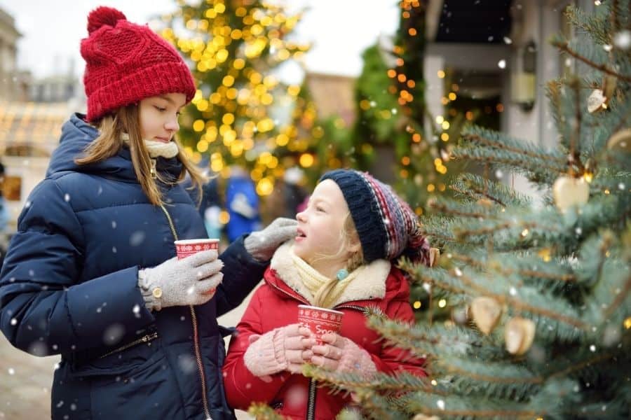 Knoxville Christmas Events represented by 2 young girls holding hot chocolate by an outdoor Christmas tree