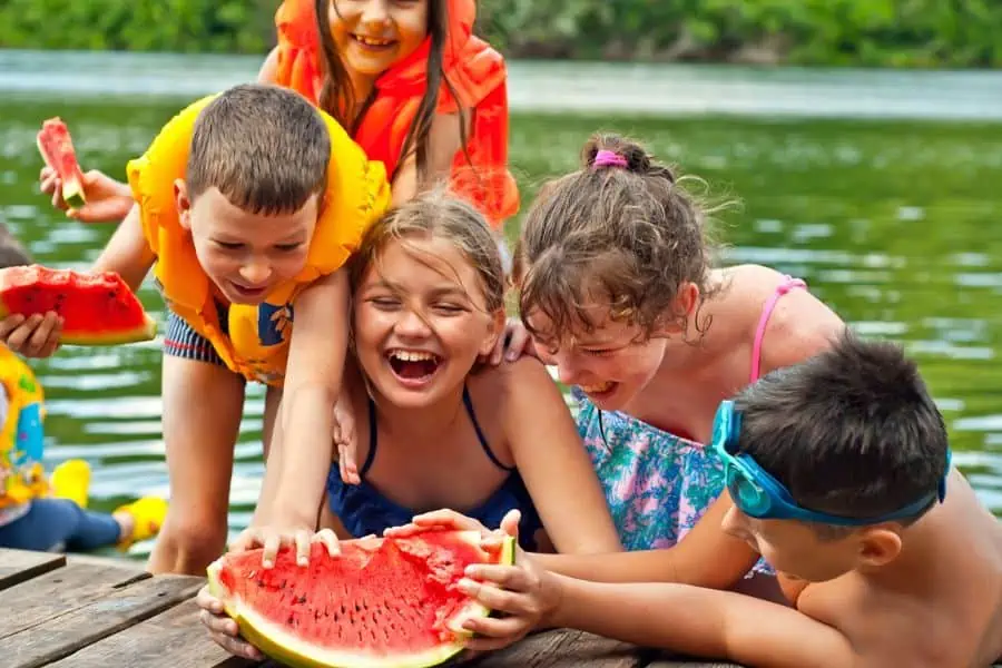 Happy group of kids laughing and eating watermelon at lake.