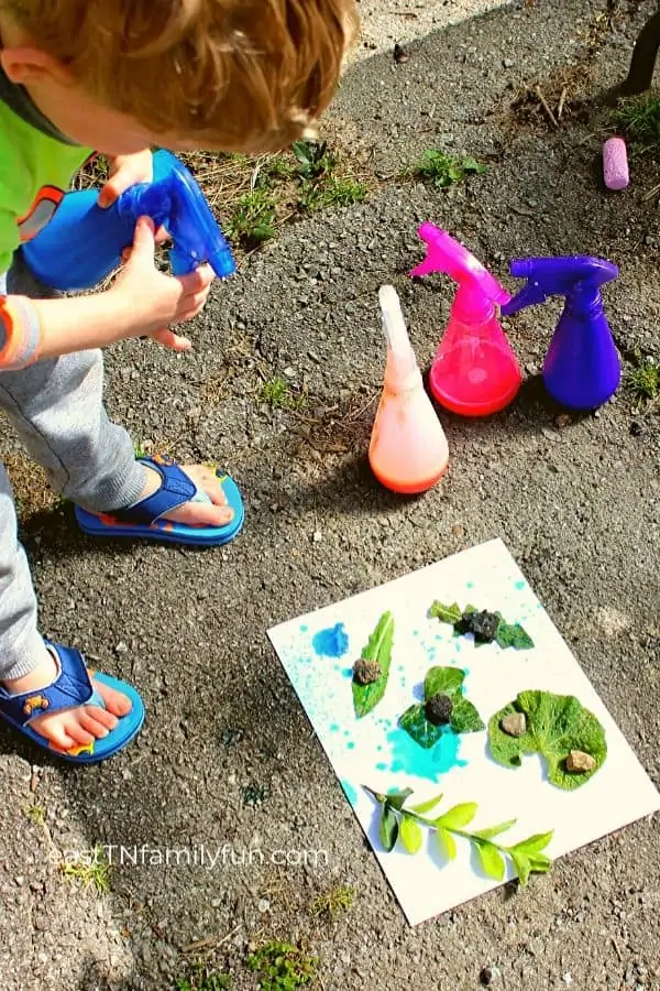 Child spraying blue color onto a white paper covered in leaves