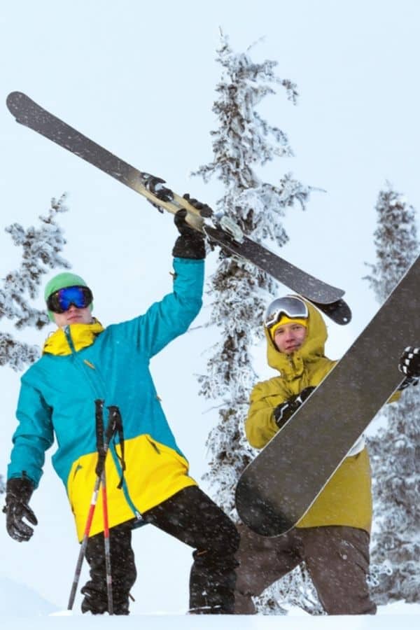 two friends hoisting their skis and snowboard overhead on the top of a mountian peak